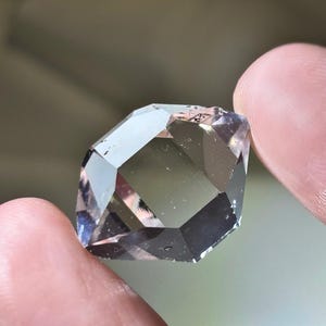 May include: A faceted, clear gemstone with a geometric shape. The gem has multiple reflective surfaces, creating a play of light and shadow. The stone is held between two fingers, showcasing its size and clarity.