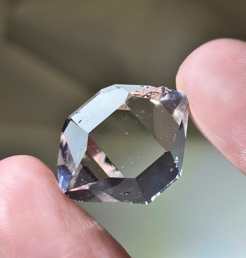 May include: A faceted, clear gemstone with a geometric shape. The gem has multiple reflective surfaces, creating a play of light and shadow. The stone is held between two fingers, showcasing its size and clarity.