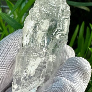 May include: A clear, raw quartz crystal specimen with a jagged, irregular surface texture and multifaceted edges. The stone is translucent, allowing light to pass through its crystalline structure, showcasing natural ridges and deep grooves.