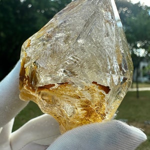 May include: A large, clear quartz crystal with a rough, natural shape. The crystal has a golden brown colour throughout, with a few small, dark brown inclusions. The crystal is being held in a white glove.