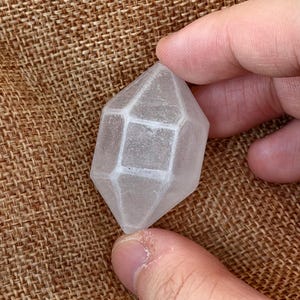 Op de afbeelding: Een helder, doorschijnend kristal met dubbele punt en een geometrische, gefacetteerde vorm. Het kristal heeft een centraal zeshoekig gedeelte dat aan beide zijden uitloopt in punten, met een glad, gepolijst oppervlak en zichtbare insluitingen.