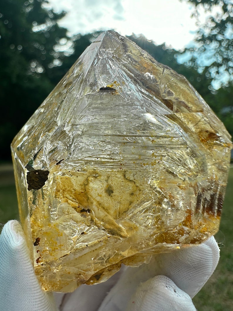 May include: A large, clear quartz crystal with a golden hue and inclusions. The crystal is faceted and has a natural, rough texture.
