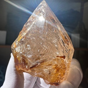 May include: A large, clear quartz crystal with a brown, natural inclusion. The crystal is faceted and has a pointed tip. It is being held in a white glove.