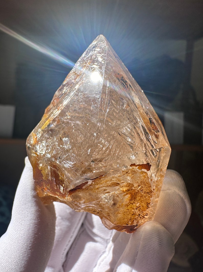 May include: A large, clear quartz crystal with a brown, natural inclusion. The crystal is faceted and has a pointed tip. It is being held in a white glove.