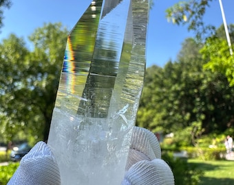 May include: A large, clear quartz crystal point held in gloved hands against a backdrop of green trees and a blue sky. The crystal has a sharp, faceted shape with internal rainbow reflections. The base of the crystal is a cloudy white.