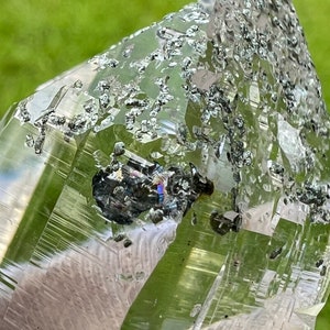 May include: A clear quartz crystal with a green hue and inclusions of black hematite. The crystal is pointed at the top and has a smooth, polished surface.