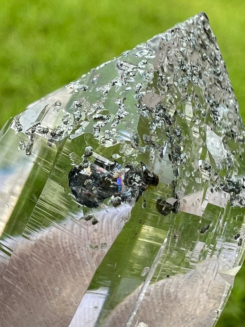 May include: A clear quartz crystal with a green hue and inclusions of black hematite. The crystal is pointed at the top and has a smooth, polished surface.
