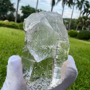 May include: A large, clear quartz crystal with a rough, natural shape. The crystal is held in a white glove and has a slight green tint.