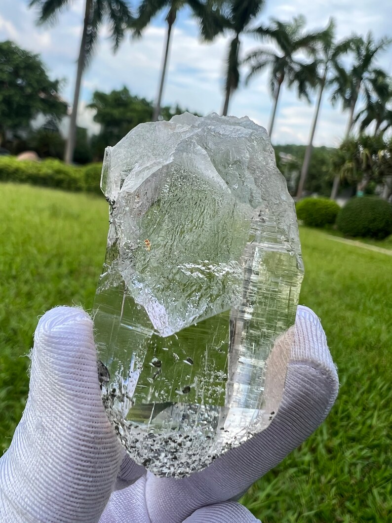 May include: A large, clear quartz crystal with a rough, natural shape. The crystal is held in a white glove and has a slight green tint.