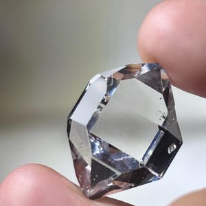 Op de afbeelding: Een heldere, gefacetteerde edelsteen met een complexe geometrische vorm. De edelsteen wordt tussen twee vingers gehouden, waarbij de reflecterende oppervlakken en de interne helderheid worden benadrukt. De steen lijkt transparant, met subtiele insluitsels.
