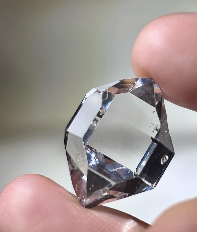 May include: A clear, faceted gemstone with a complex geometric shape. The gem is held between two fingers, showcasing its multiple reflective surfaces and internal clarity. The stone appears to be transparent, with subtle inclusions visible within its structure.