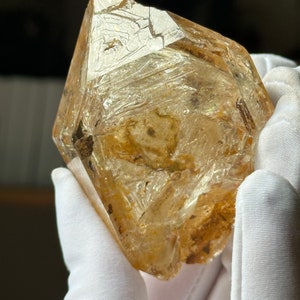 May include: A large, clear, faceted quartz crystal with a golden hue. The crystal has a rough, textured surface and is covered in small, white inclusions. The crystal is held in a pair of white gloves.