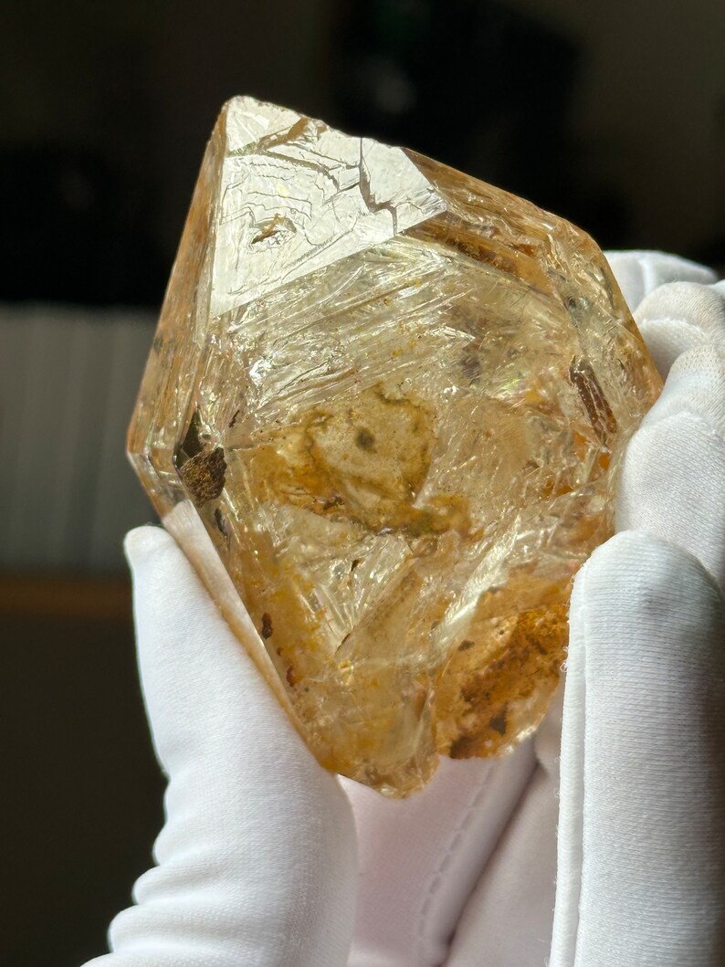 May include: A large, clear, faceted quartz crystal with a golden hue. The crystal has a rough, textured surface and is covered in small, white inclusions. The crystal is held in a pair of white gloves.