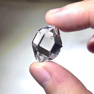 May include: A clear, faceted gemstone with a geometric shape. The gem is held between two fingers, showcasing its transparent quality and internal reflections. The stone appears to be a diamond or a similar crystal.