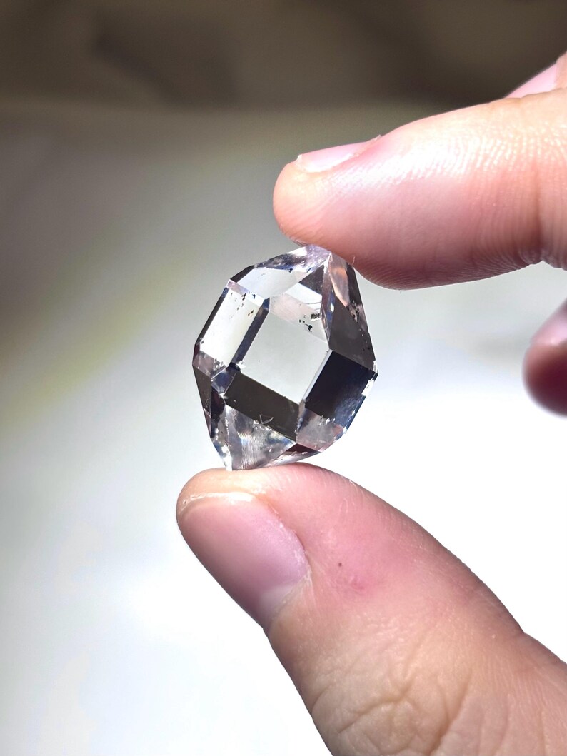 May include: A clear, faceted gemstone with a geometric shape. The gem is held between two fingers, showcasing its transparent quality and internal reflections. The stone appears to be a diamond or a similar crystal.