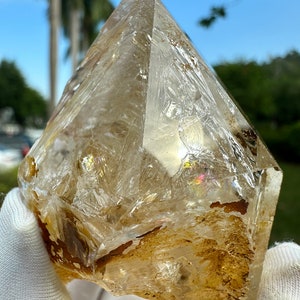May include: A large, clear quartz crystal with a golden brown hue. The crystal has a pointed tip and a rough, natural surface. It is a beautiful example of a natural crystal.