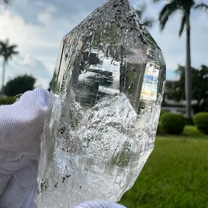 May include: A large, clear quartz crystal with a pointed tip and many facets. The crystal is held in a white gloved hand and has a cloudy, milky appearance.