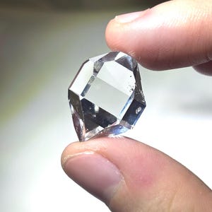 May include: A clear, faceted gemstone with multiple reflective surfaces. The gem is held between two fingers, showcasing its geometric shape and clarity. The stone appears to be a diamond or a similar crystal, reflecting light and creating a sparkling effect.