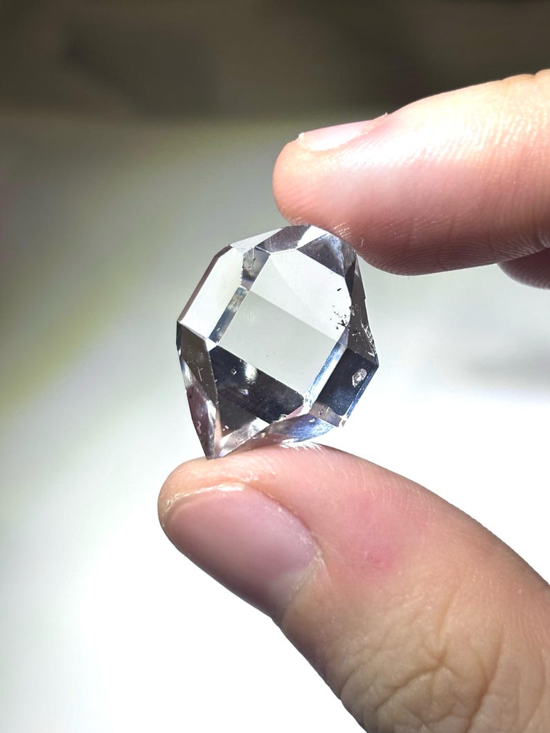 May include: A clear, faceted gemstone with multiple reflective surfaces. The gem is held between two fingers, showcasing its geometric shape and clarity. The stone appears to be a diamond or a similar crystal, reflecting light and creating a sparkling effect.