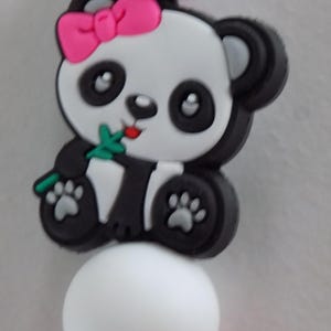 Puede incluir: Clip para chupete con temática de oso panda, con una combinación de colores blanco, negro y rosa. El panda tiene un lazo rosa y sostiene una hoja verde. El clip tiene una cuenta blanca y una cuenta rosa.