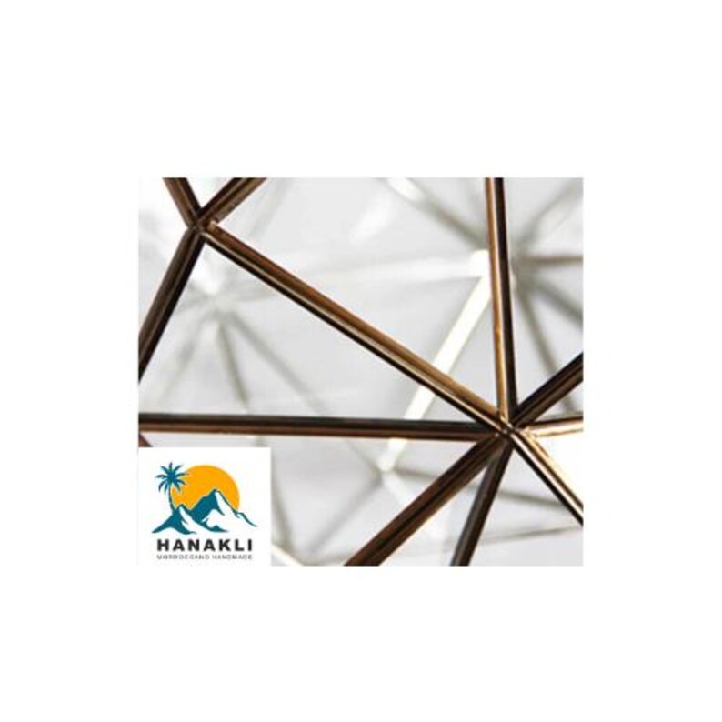 May include: Close-up of a geometric metal frame with a white background. The frame is made of thin, gold-colored metal bars that intersect at various angles.