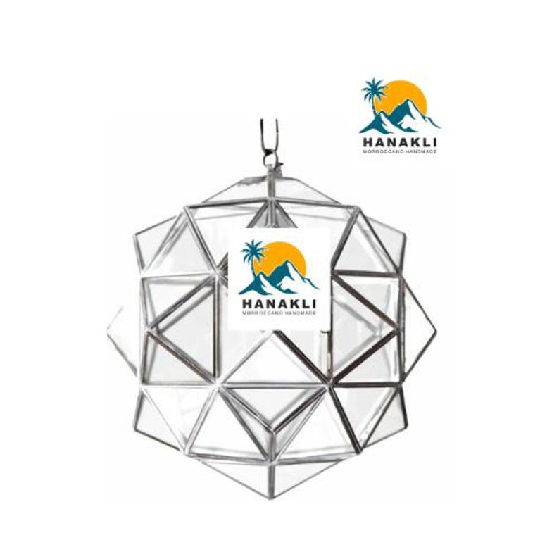 May include: A silver geometric terrarium with a clear glass design. The terrarium is shaped like a sphere and has a small opening at the top. The terrarium is sitting on a white surface. The text "HANAKLI MOROCCO HANDMADE" is visible in the image.