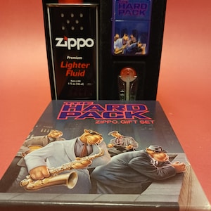 May include: A Zippo lighter gift set featuring a purple lighter with a band of musicians and the text "The Hard Pack" on the front. The set also includes a bottle of Zippo lighter fluid and a black box with the text "The Hard Pack Zippo Gift Set" on the front.