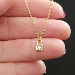 Puede incluir: Un collar de oro con un pequeño colgante de candado con una piedra de cristal transparente.