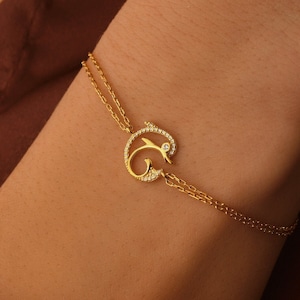 Könnte beinhalten: Ein goldenes Kettenarmband mit einem zarten, verzierten Anhänger, der einen stilisierten Buchstaben 'G' mit funkelnden, klaren Steinen zeigt.