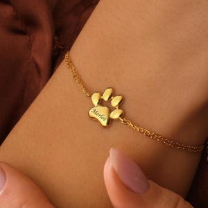 May include: A gold-coloured bracelet with a paw print charm engraved with the name "Merlot". The charm is attached to a fine chain. The bracelet is displayed on a wrist.