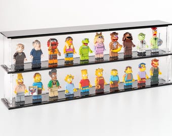 May include: A clear acrylic display case with two shelves, showcasing a collection of miniature figurines. The figurines include characters from The Muppets and The Simpsons, with vibrant colors and detailed designs. The case has a black base and top.