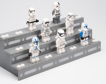 May include: A three-tiered gray plastic display stand featuring white circular studs on each level, holding seven assorted Star Wars-themed minifigures with white, black, and blue armor details, including various helmet designs and printed torso patterns.