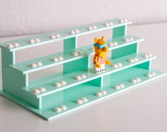 May include: A tiered, mint-green display stand with multiple levels, each adorned with white, circular studs. A yellow Lego figure with sunglasses and a backpack stands on the middle tier. The stand is designed to showcase small collectibles.