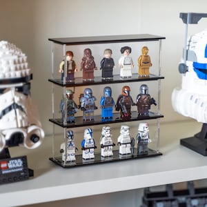 Peut inclure: Un présentoir en acrylique transparent à trois étagères, présentant diverses figurines Lego Star Wars. Le présentoir a un dessus et une base noirs et est rempli de personnages comme Dark Vador, Boba Fett et des Stormtroopers. D'autres modèles Lego Star Wars sont visibles en arrière-plan.
