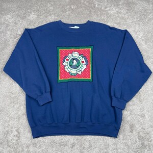 May include: A navy blue long-sleeve sweatshirt featuring a central square graphic. The graphic has a red background with white dots, a green border, and a circular design in the middle depicting a snowman, green trees, and white houses.