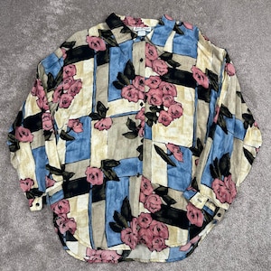 May include: A long-sleeve button-down shirt featuring a geometric grid pattern in shades of blue, cream, and black, overlaid with a floral print of pink roses and dark green leaves. The shirt has a standard collar and a full-length front button closure.
