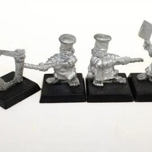 Pode incluir: Quatro figuras de chefes em miniatura prateadas em várias poses. Uma figura puxa uma corda, enquanto outras seguram um cutelo ou apontam. Cada figura é montada em uma base quadrada preta, em um fundo branco.