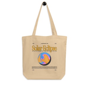 Op de afbeelding: Een beige tote bag met een grafisch ontwerp van een zonsverduistering. De tekst "I survived the Solar Eclipse" is in geel op de tas gedrukt. De zonsverduistering is afgebeeld als een draaiende oranje, roze en blauwe bol.