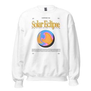 Op de afbeelding: Witte sweatshirt met de tekst "I Survived the Solar Eclipse" in geel en zwart. Daaronder een afbeelding van een kleurrijke planeet met blauwe, oranje en roze wervelingen. De sweatshirt heeft een ronde hals en lange mouwen.