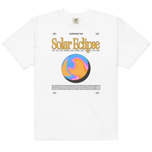Op de afbeelding: Wit T-shirt met een grafische print van een planeet met een draaiende print van blauw, roze en oranje. De tekst "I Survived The Solar Eclipse" is in geel boven de planeet gedrukt.