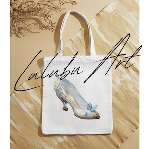 May include: White tote bag featuring a watercolor illustration of a glass slipper with blue butterfly accents. The bag has the text "Lalabu Art" in a handwritten font.