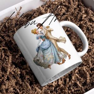 May include: White ceramic mug featuring a romantic illustration of a couple dancing in formal attire. The woman wears a blue gown, and the man is in a gold coat. The mug is inside a white box with brown paper filler. The text "Calubu Art" is written on the mug.