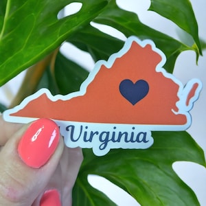 May include: A sticker featuring the outline of the state of Virginia in orange, with a navy blue heart in the center. The word "Virginia" is written in a light blue font below the state outline. The sticker is held in front of a green leaf.