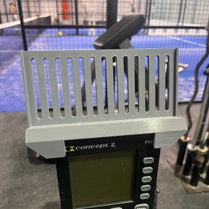 May include: A Concept 2 PM rowing machine monitor with a gray plastic shield. The monitor has a black screen and several buttons. The shield has vertical slots. The machine is in a gym setting.