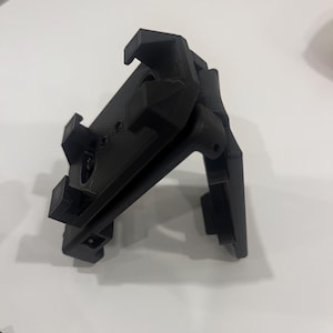 Pode incluir: Um dispositivo preto impresso em 3D com múltiplos componentes retangulares e angulares. O dispositivo tem uma base retangular com vários braços e clipes salientes. O objeto é projetado com uma série de orifícios e ranhuras.