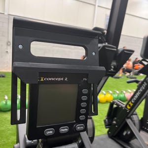Op de afbeelding: Zwarte Concept 2 roeimachine met een digitaal display met eenheden, display en menu-opties. De machine heeft een zwart frame en staat in een sportschool met andere fitnessapparatuur.