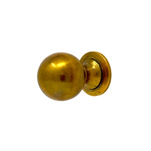 May include: A gold-toned round cabinet knob with a smooth, shiny surface. The knob has a circular base with a raised edge.