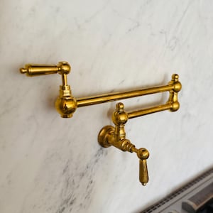 May include: A gold-toned kitchen faucet with a double-jointed arm and a single handle. The faucet is mounted to a white marble wall.