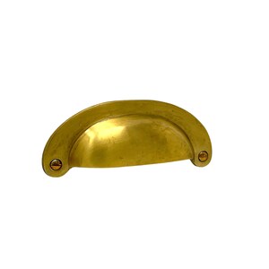 May include: A solid brass cup pull with a half-moon shape. The hardware has a warm, golden hue and is secured with two screws. This cabinet pull is a classic design element for furniture.