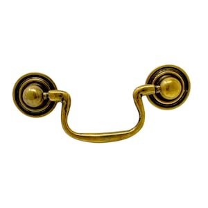May include: A gold-toned metal cabinet pull with a rounded, decorative design. The pull has a simple, curved shape and two round knobs on either end.